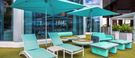 Zona relax esterna moderna con poltrone e ombrelloni color acquamarina, circondata da pareti vetrate e un prato.