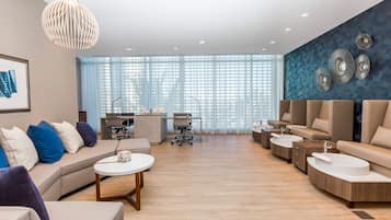 A modern hotel lobby with a sofa, armchairs, a desk, and a large window with blinds.