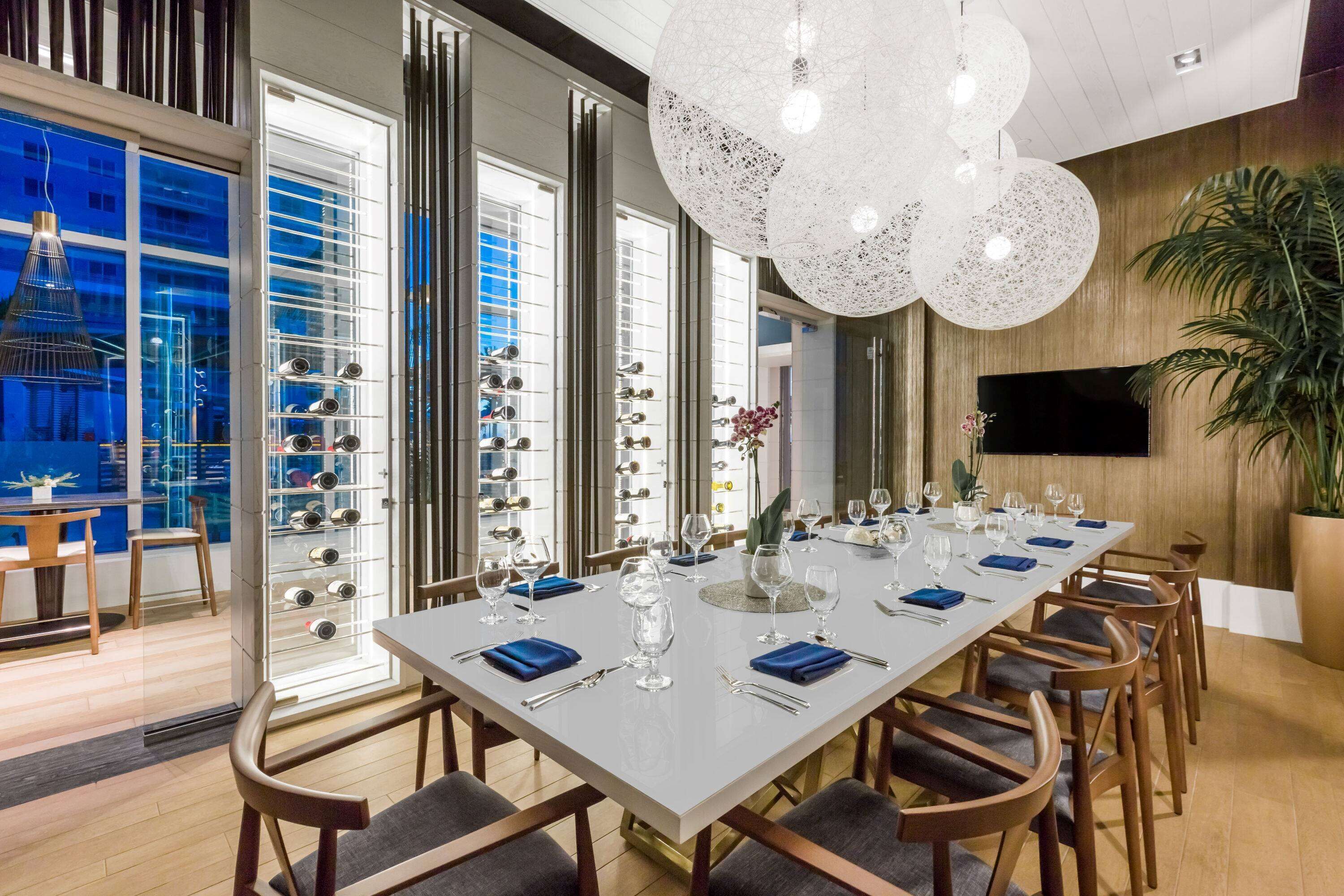 A modern dining area with a long table set for a meal, large windows, and a large white chandelier.