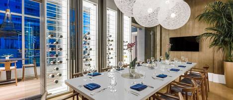 A modern dining area with a long table set for a meal, large windows, and a large white chandelier.