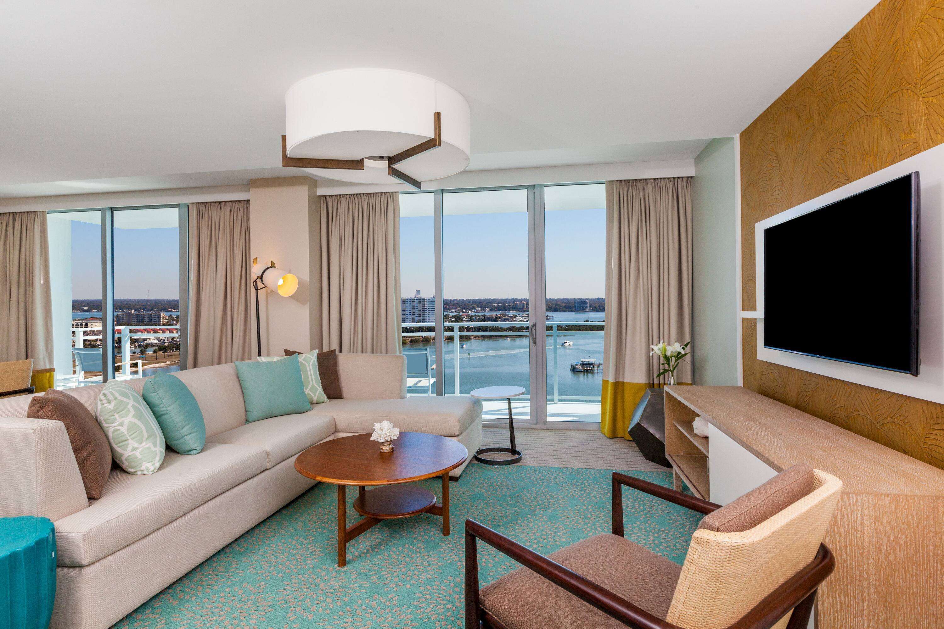 A hotel room with a large sofa, a coffee table, a TV, and a view of a body of water through large windows.