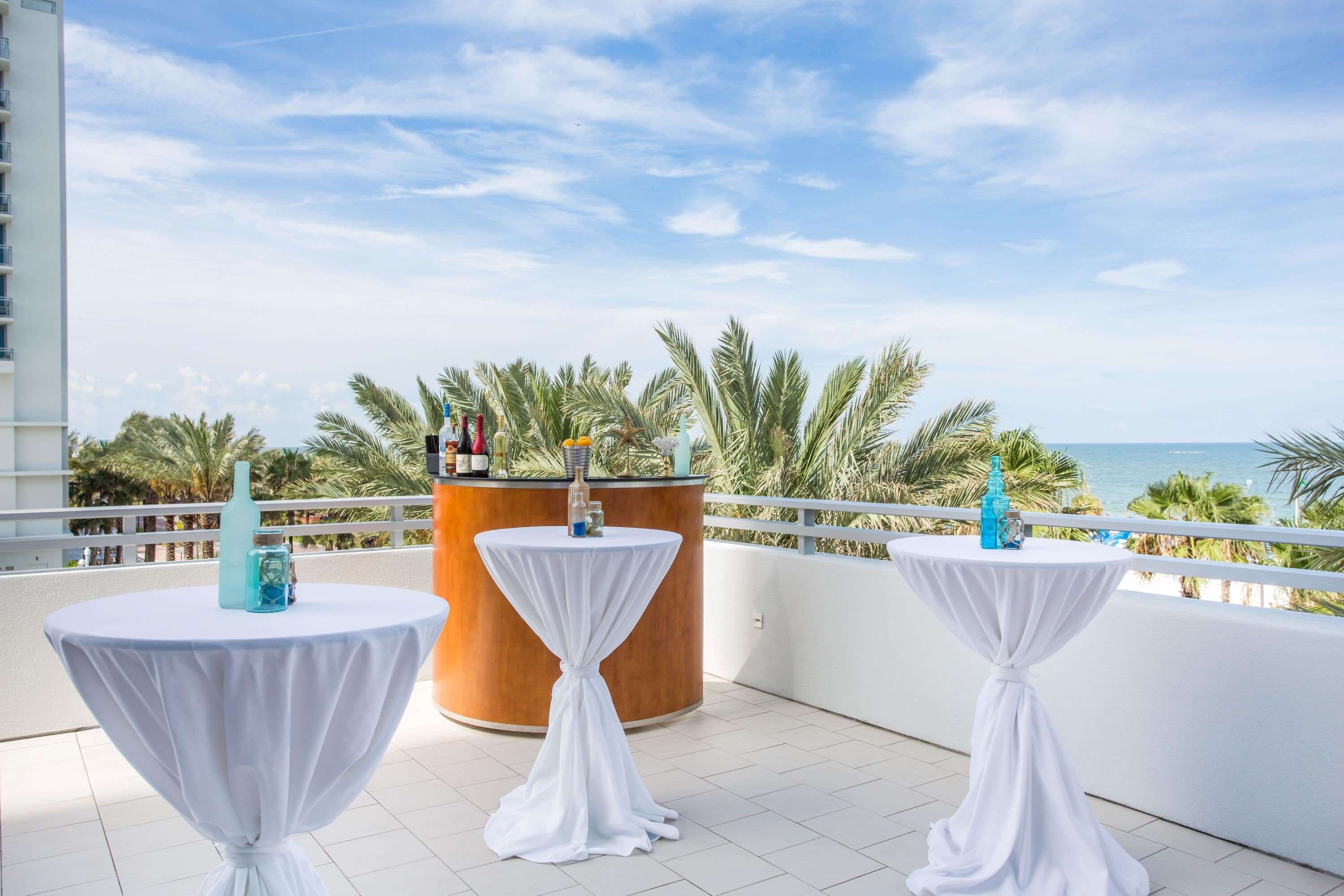 A balcony with three round tables covered with white cloths, a wooden bar, and a view of the sea and palm trees.