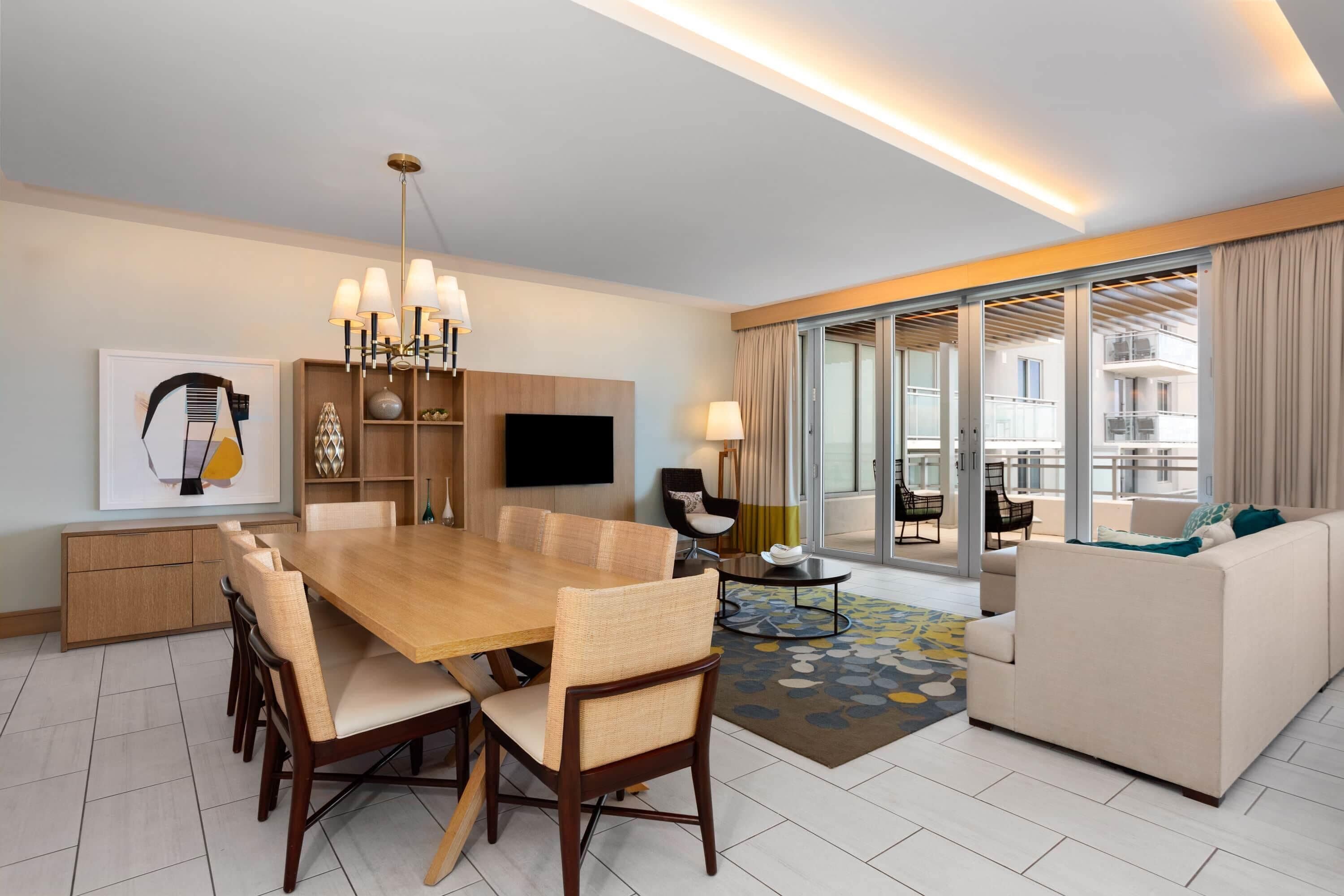 A modern hotel room with a dining area, a sofa, a TV, and a balcony with a view.