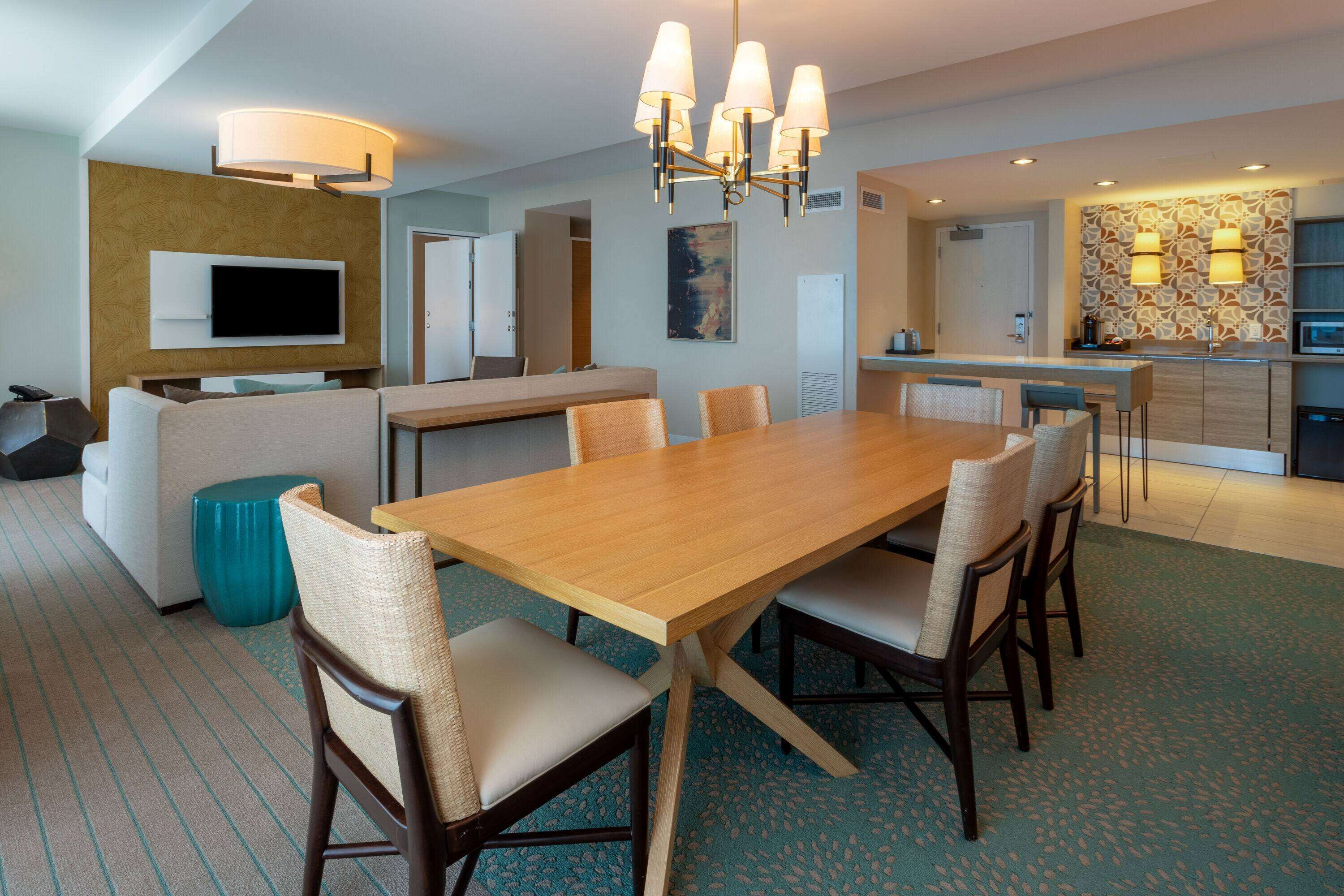 A modern hotel room with a dining area, a sofa set, a TV mounted on the wall, and a kitchen area with a microwave.