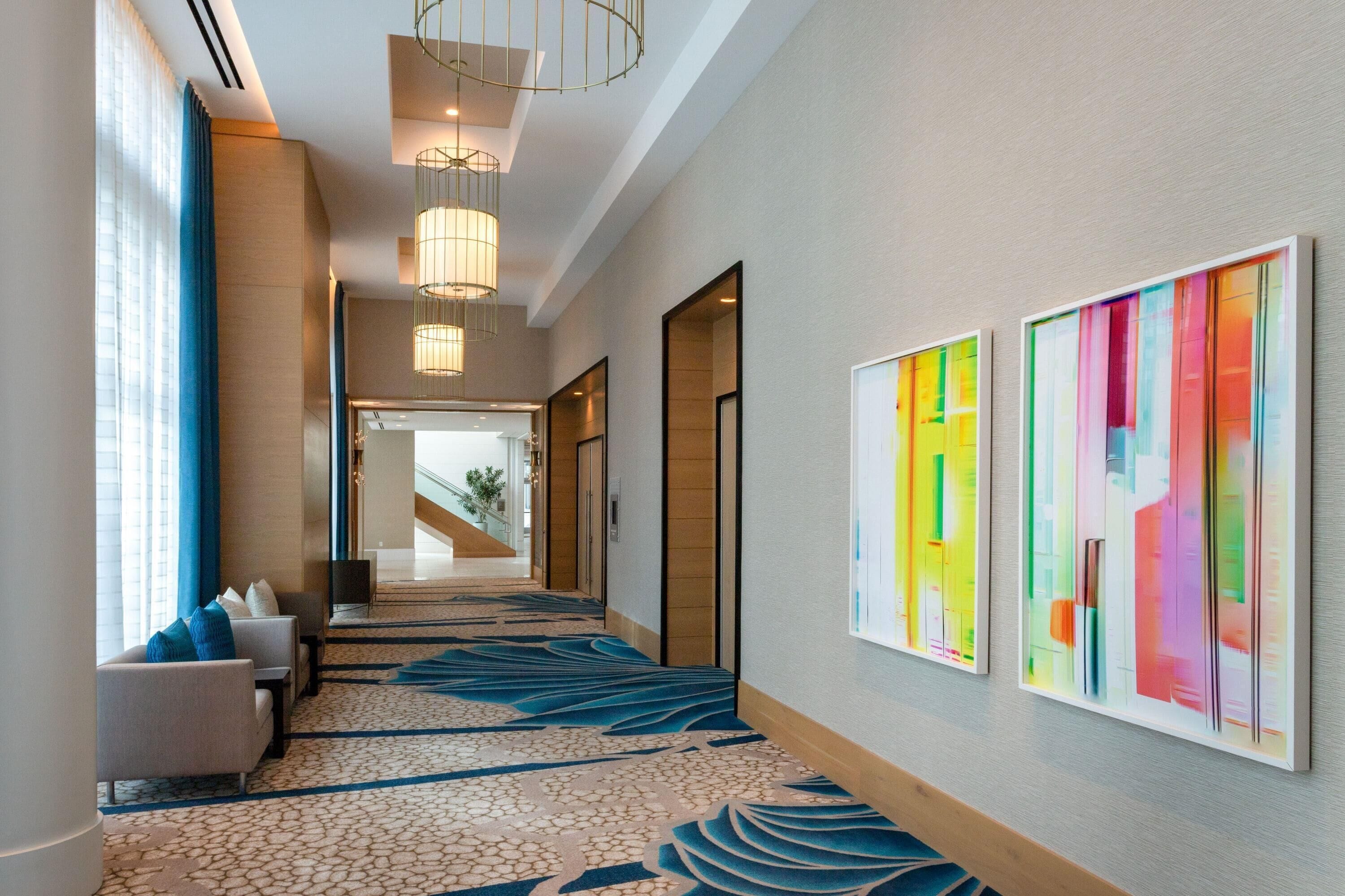 A hotel hallway with a patterned carpet, seating area with a sofa and armchairs, and two colorful abstract paintings on the wall.