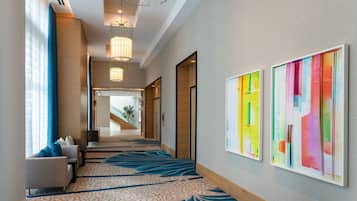 Un pasillo de hotel con alfombra estampada, una zona de estar con sofá y sillones, y dos cuadros abstractos coloridos en la pared.