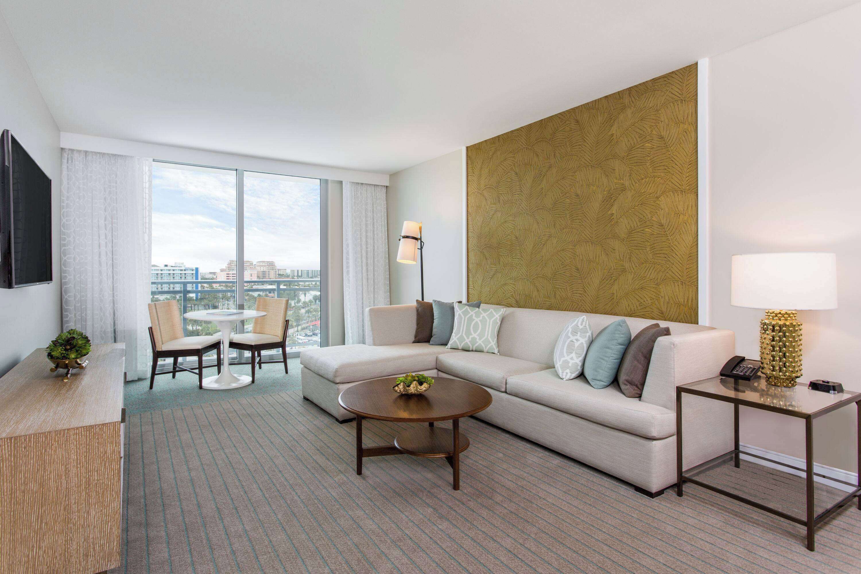 Una habitación de hotel moderna con sofá, una mesa de centro, un escritorio y una silla. Se aprecia un amplio ventanal con vistas a la ciudad.