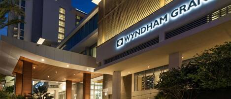 A modern hotel entrance with a prominent 'WYNDHAM GRAND' sign, a covered walkway, and landscaped areas.