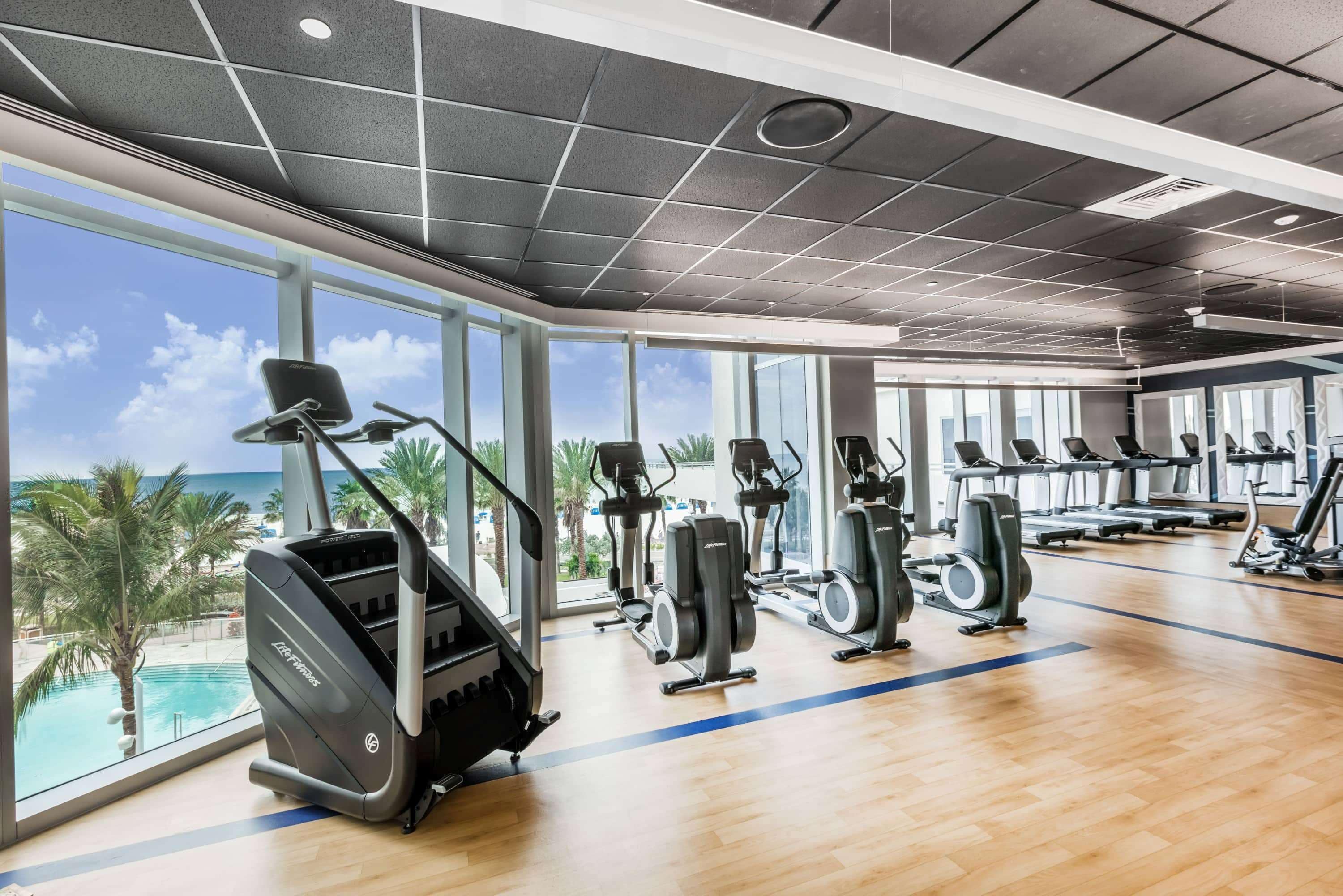 Un gimnasio con equipo de cardio y amplias ventanas con vistas a una piscina y palmeras.