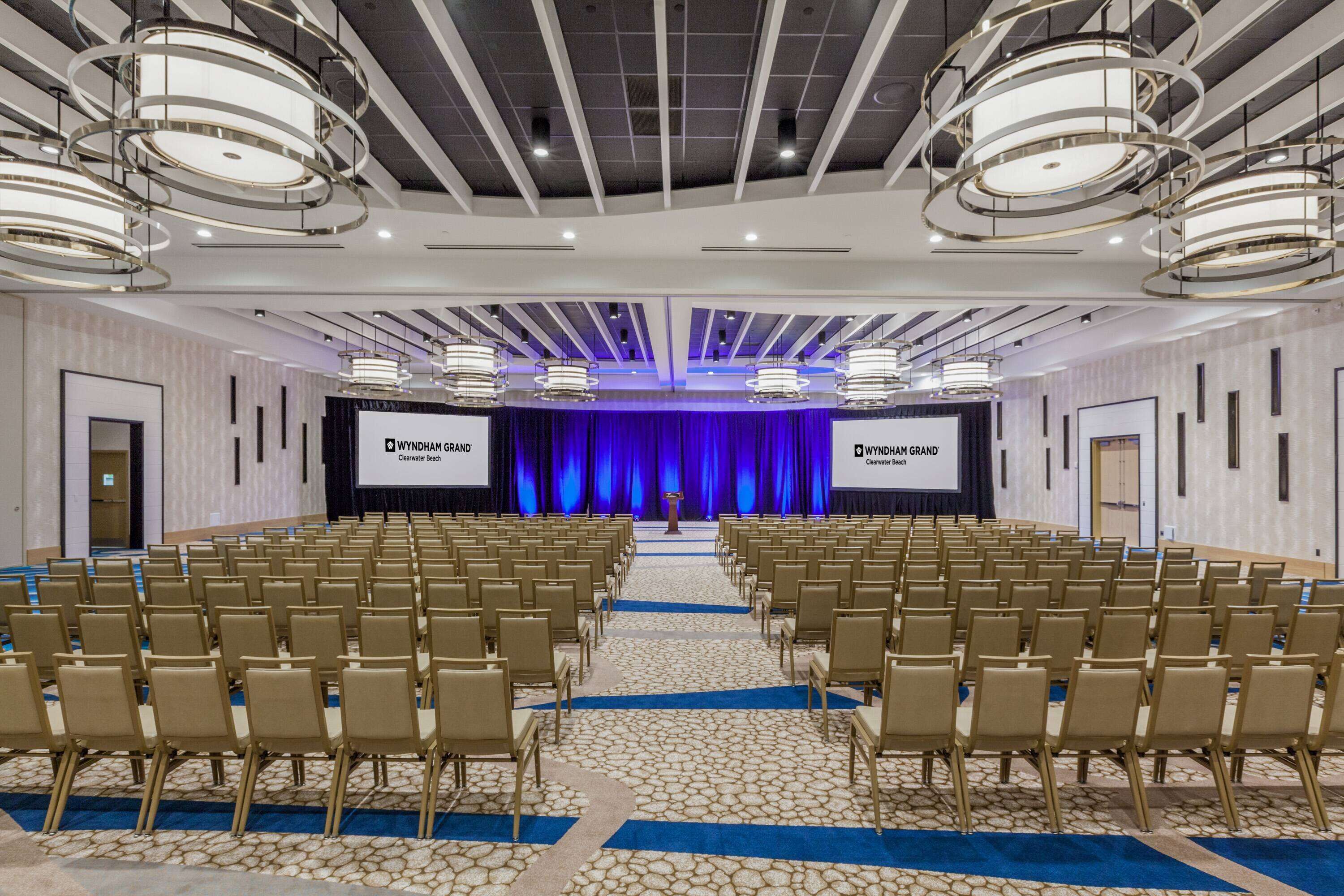 A conference room with rows of chairs, a stage with a blue curtain, and overhead lighting fixtures.
