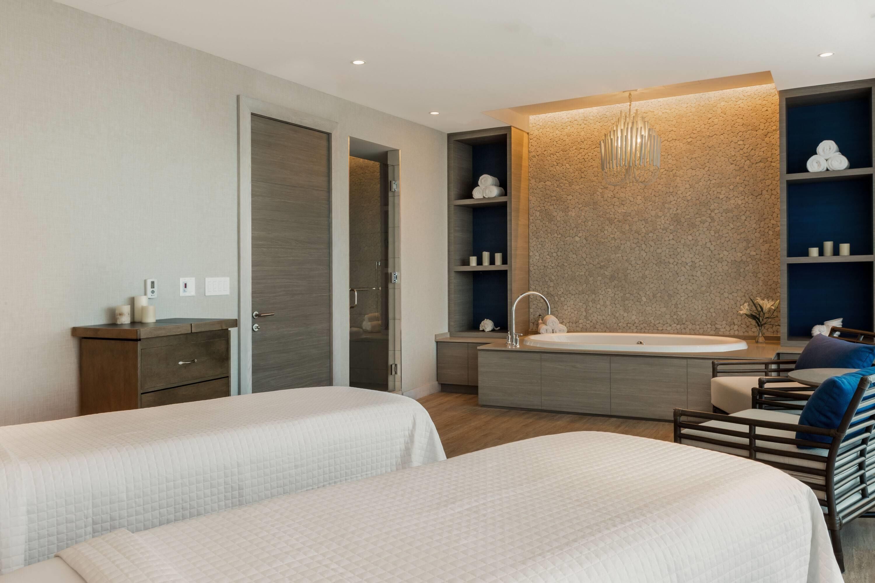 A modern hotel room with two beds, a large bathtub, a vanity with shelves, and a seating area with a chair and ottoman.