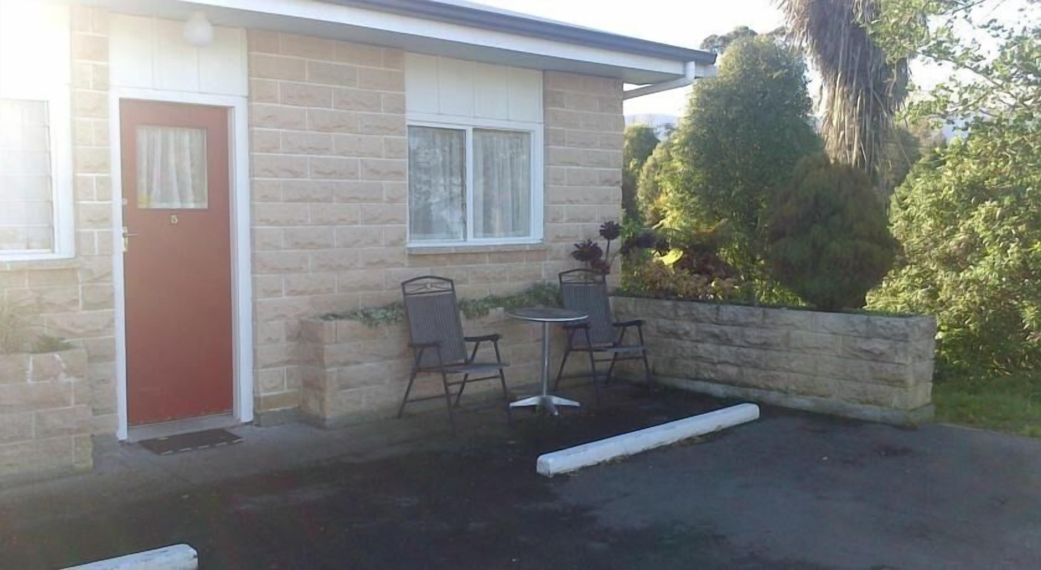 A motel with a brick exterior, a red door, and a small patio with two metal chairs and a table.