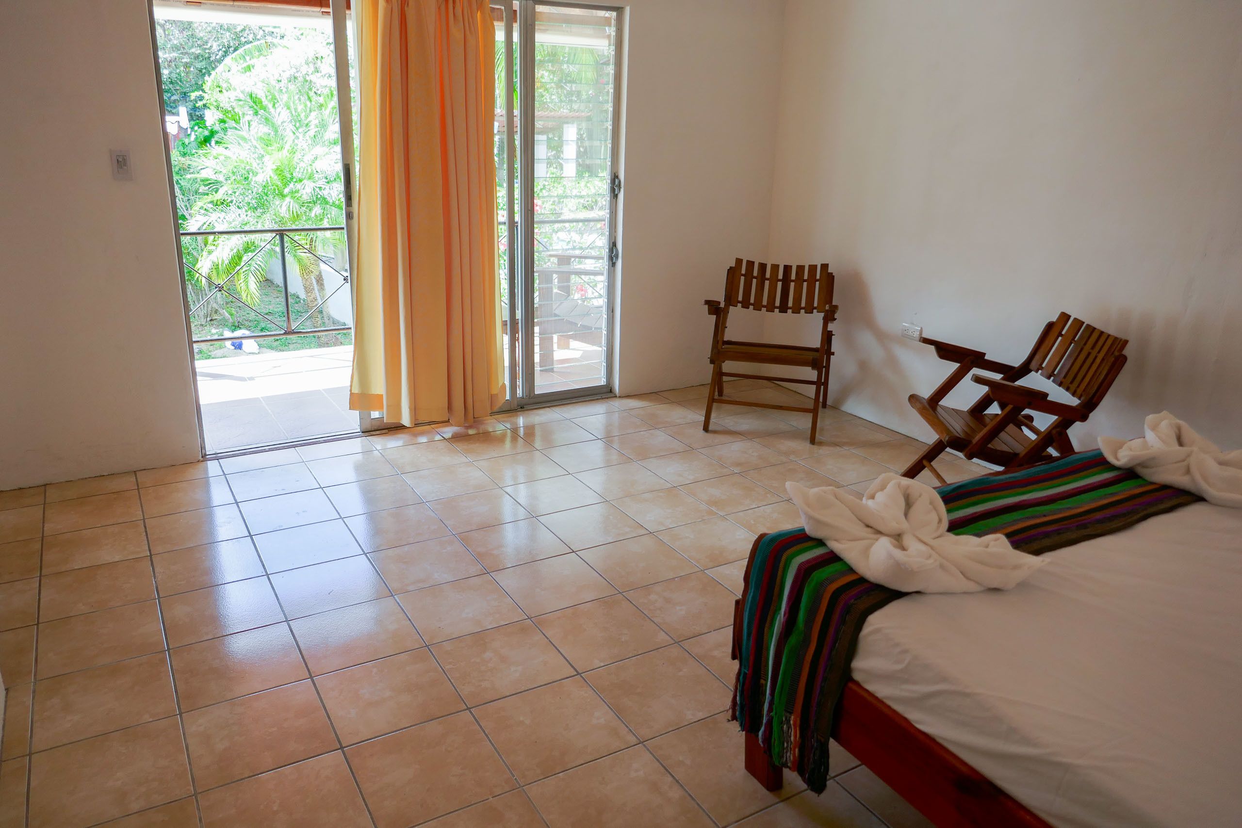 A hotel room with a bed, two wooden chairs, a balcony with curtains, and a tiled floor.
