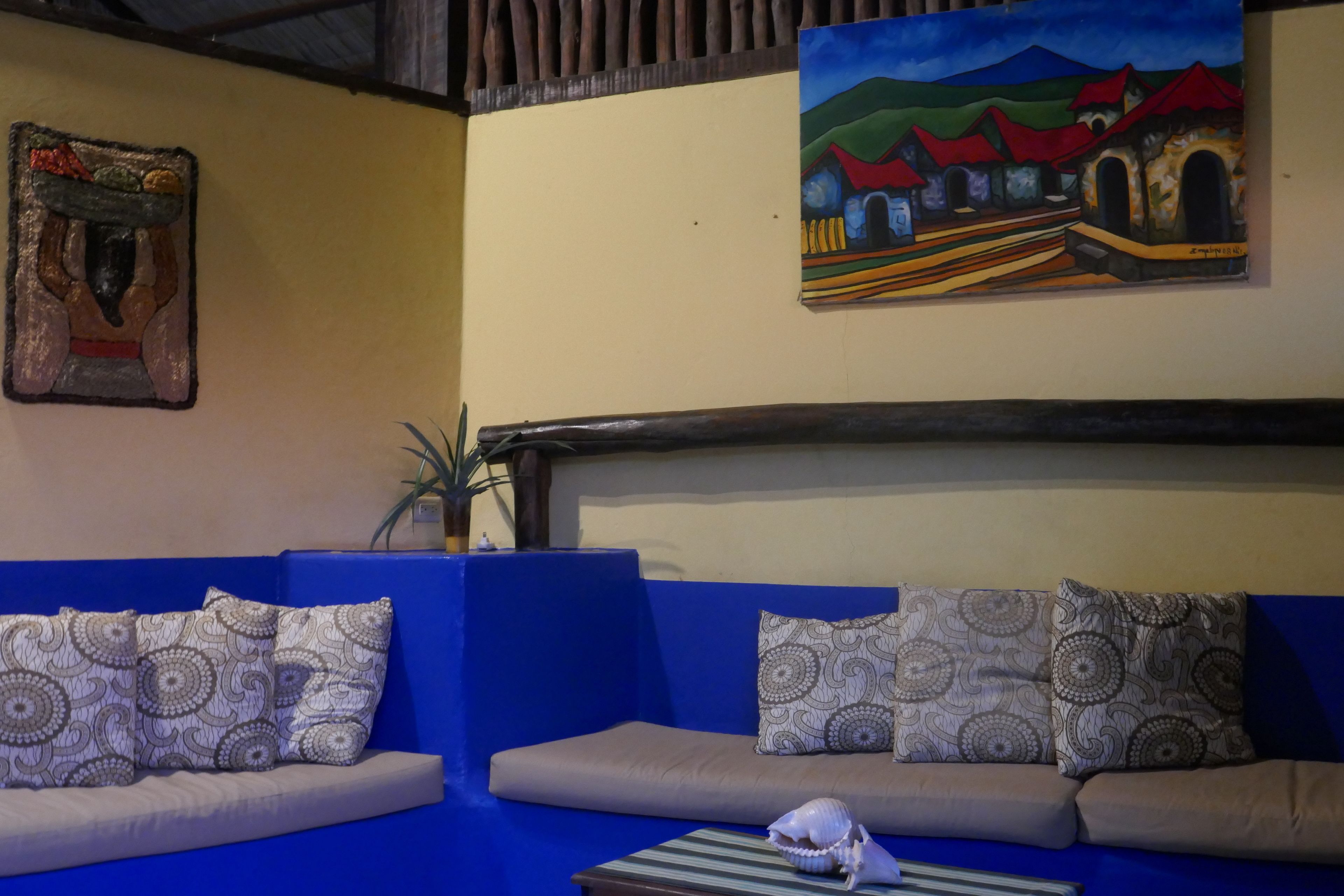 A room with two sofas, one blue and one beige, with patterned cushions. There is a painting on the wall depicting a landscape with red roofs.