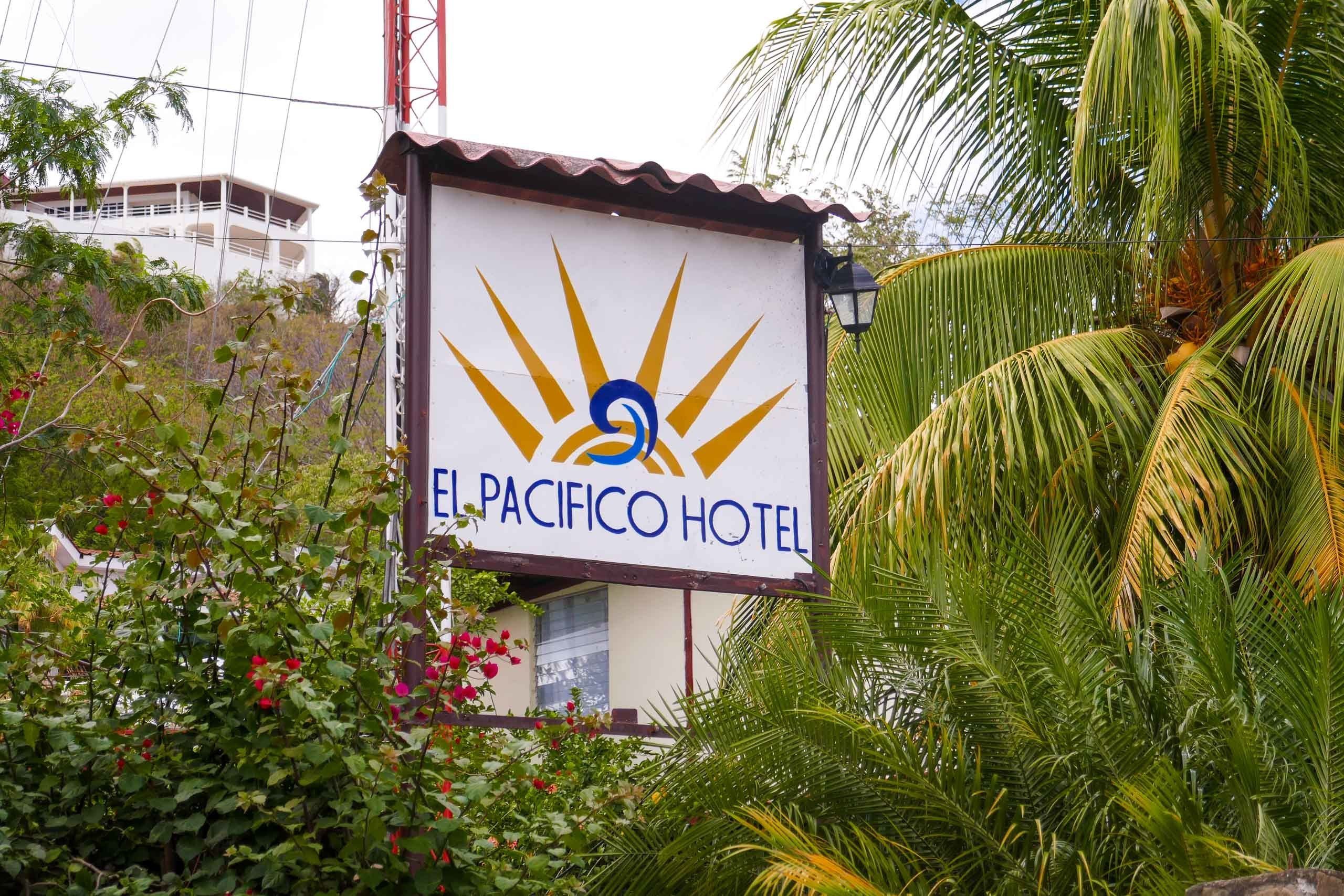 A sign for El Pacifico Hotel with a sun and wave logo, surrounded by lush greenery.