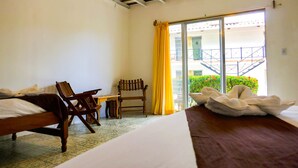 Een hotelkamer met een bed, een houten stoel, een klein tafeltje en uitzicht op een balkon met gele gordijnen.