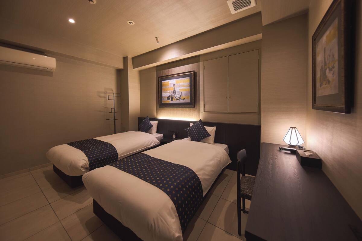 Une chambre d’hôtel avec deux lits, un bureau, une chaise, une lampe et un tableau encadré au mur.