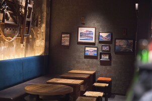 A dimly lit room with wooden tables and benches, framed artwork on the wall, and a blue cushioned bench along the wall.