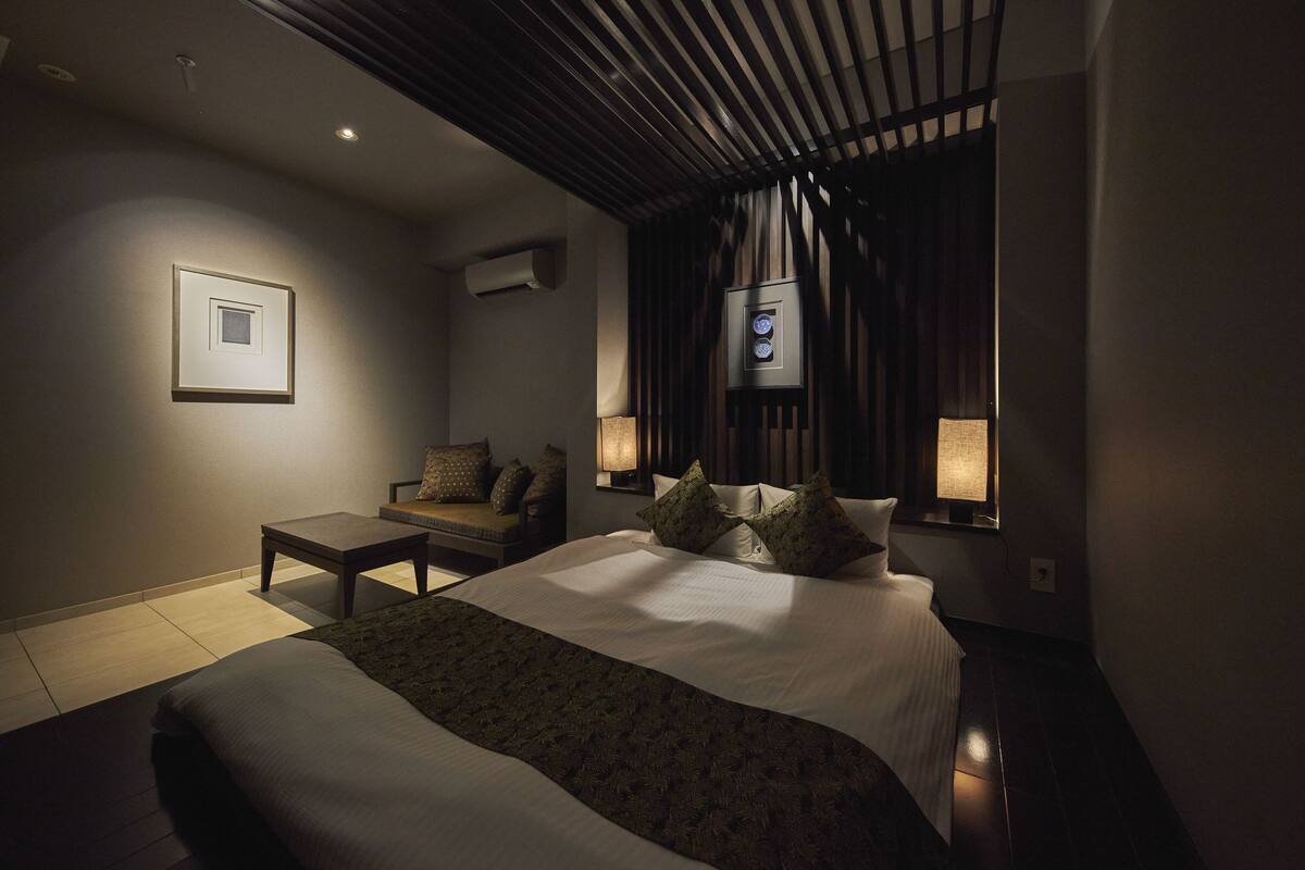 Une chambre d’hôtel moderne avec un grand lit, un petit canapé, une table de chevet et un téléviseur fixé au mur.