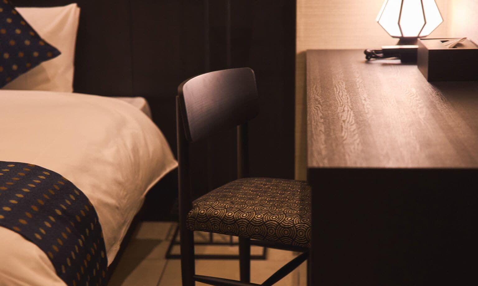Quarto de hotel com cama, poltrona, escrivaninha e abajur.