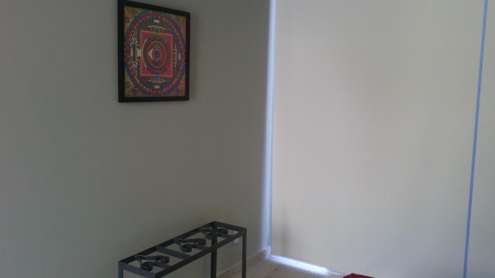 Una habitación con una obra de arte enmarcada en la pared, una pequeña mesa con estructura metálica y una esquina con pared blanca y una línea azul que la marca.