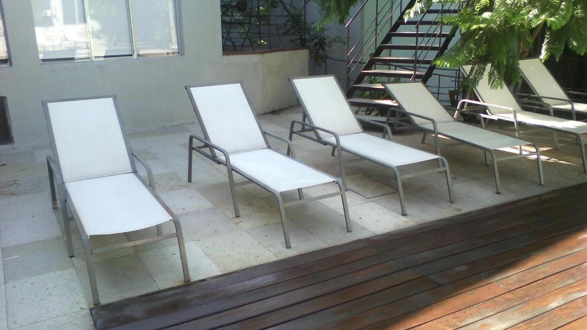 A row of white lounge chairs with metal frames, arranged on a concrete patio with a wooden deck in the foreground.