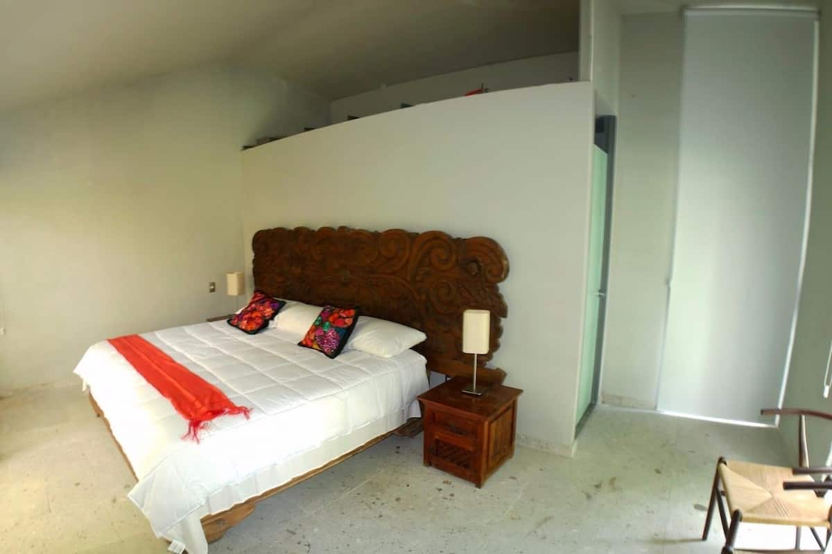 Un dormitorio con cabecera de madera, colcha blanca, dos almohadas coloridas, una mesita de noche con lámpara y una silla.