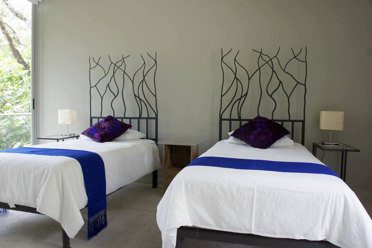 Dos camas con ropa blanca y detalles azules, un diseño decorativo de metal en la pared, y una ventana con vistas a la vegetación.