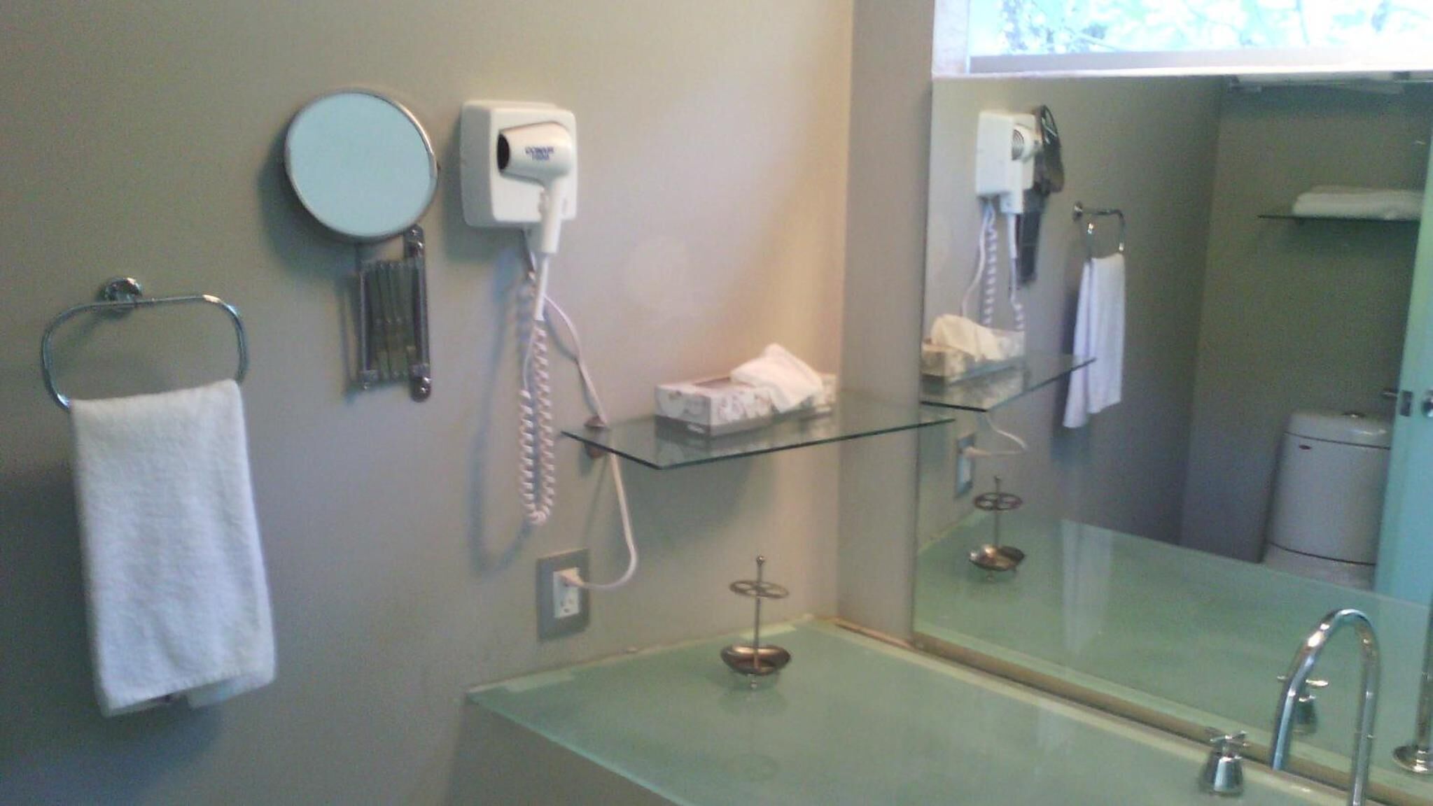 Un baño con repisa de vidrio, toallero, secador de pelo y espejo.