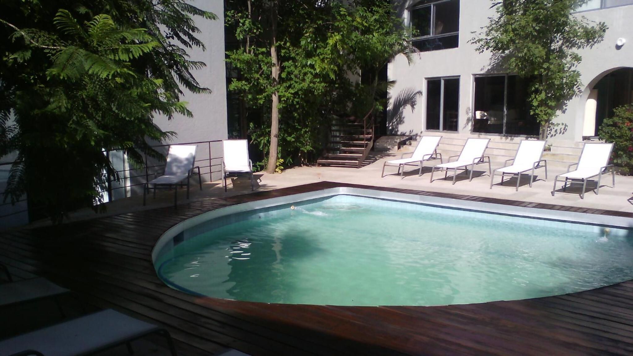 A swimming pool with a wooden deck, surrounded by white lounge chairs and a staircase leading to a building.