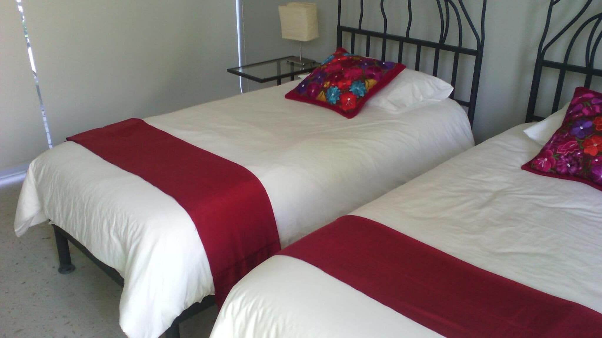 Dos camas con ropa de cama blanca y roja, un cabecero de metal y una lámpara de noche.
