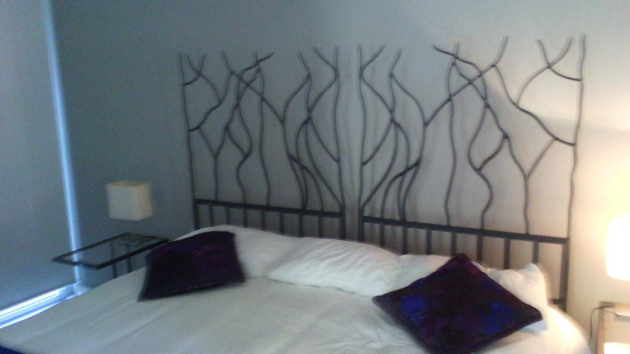 Een bed met witte lakens en twee paarse kussens, een metalen hoofdbord met een ontwerp dat aan een boom doet denken, een nachtkastje met een lamp en een bijzettafeltje met een lamp.