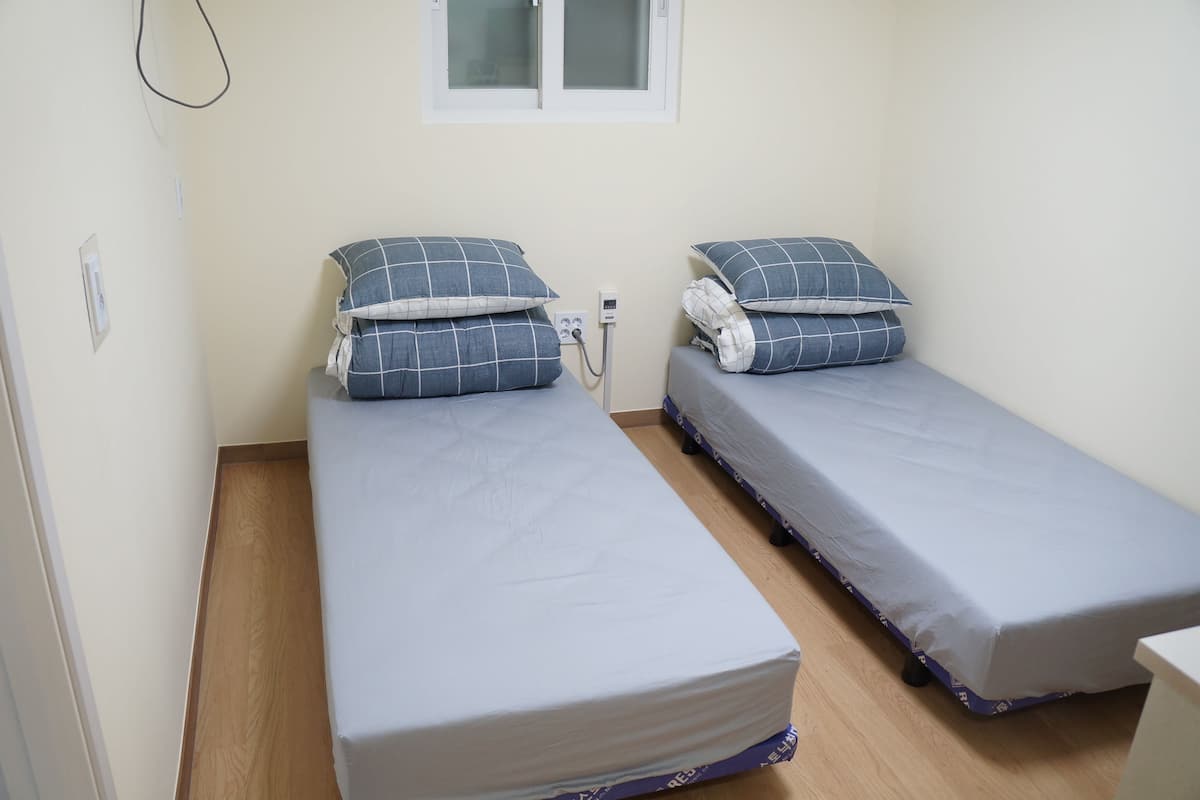 A small, single room with two single beds, each with a blue and white checkered pillow and a gray mattress. There is a window above the beds, and a power outlet is visible on the wall.