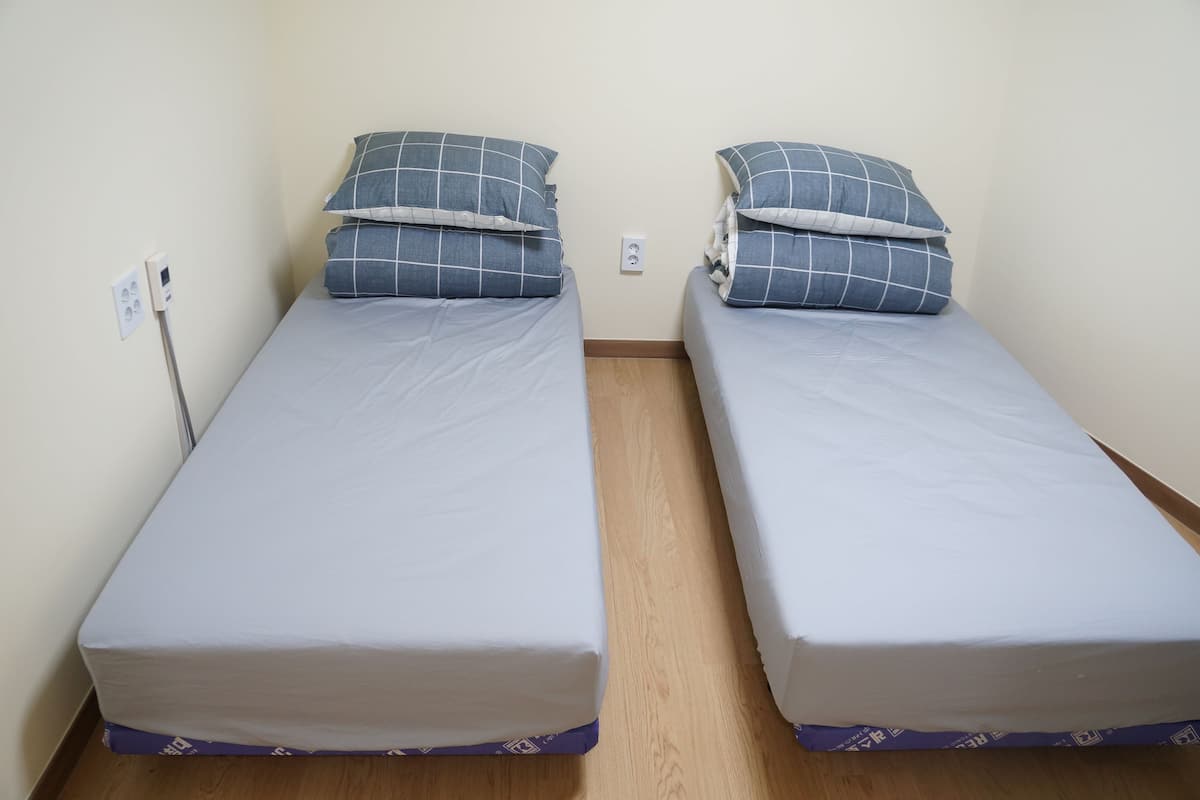 Two single beds with matching blue and white bedding, a wooden floor, and a plain wall with a power outlet.
