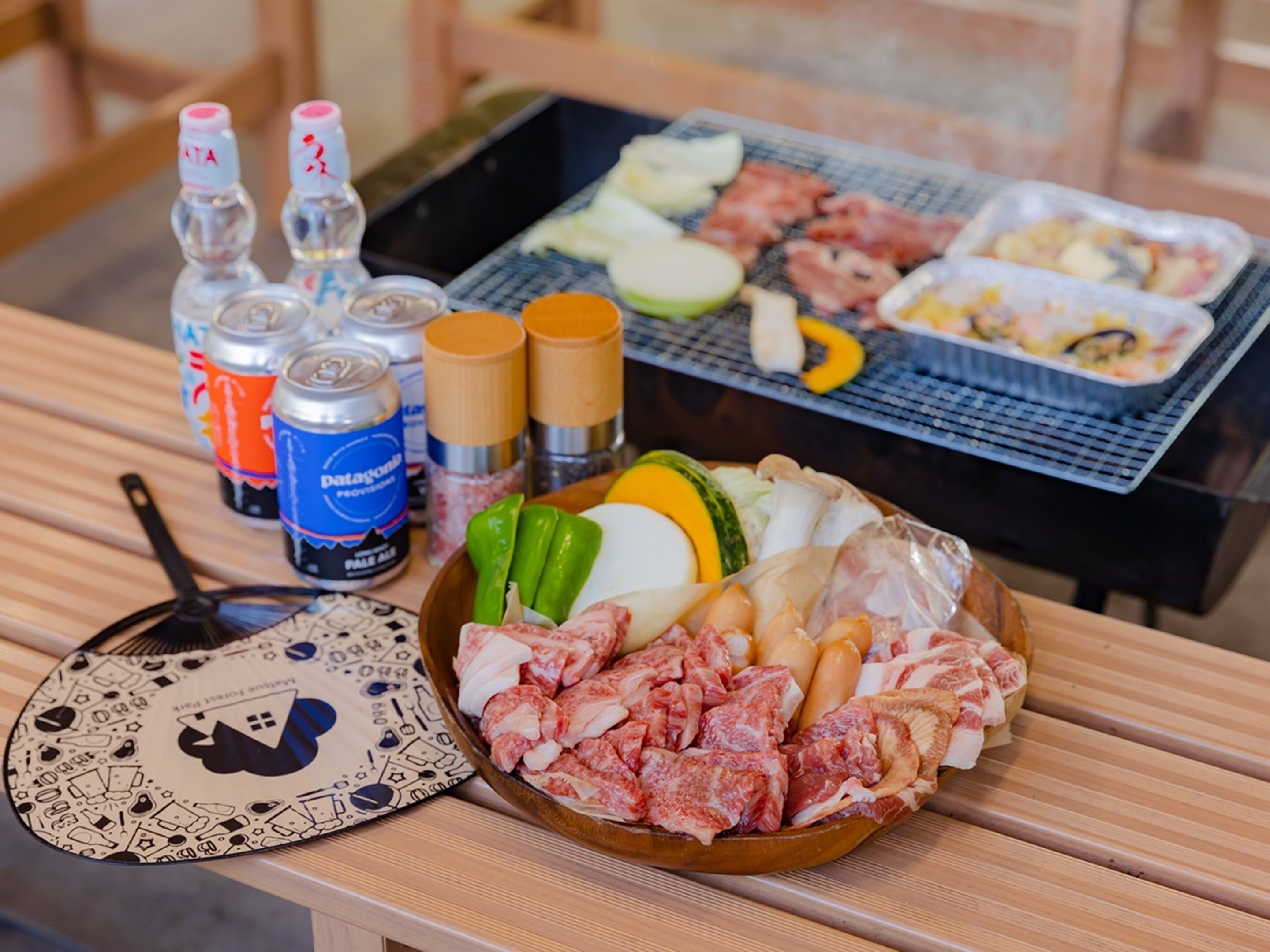A table with a variety of food items, including meat, vegetables, and condiments, along with cans of beverages and a patterned coaster.