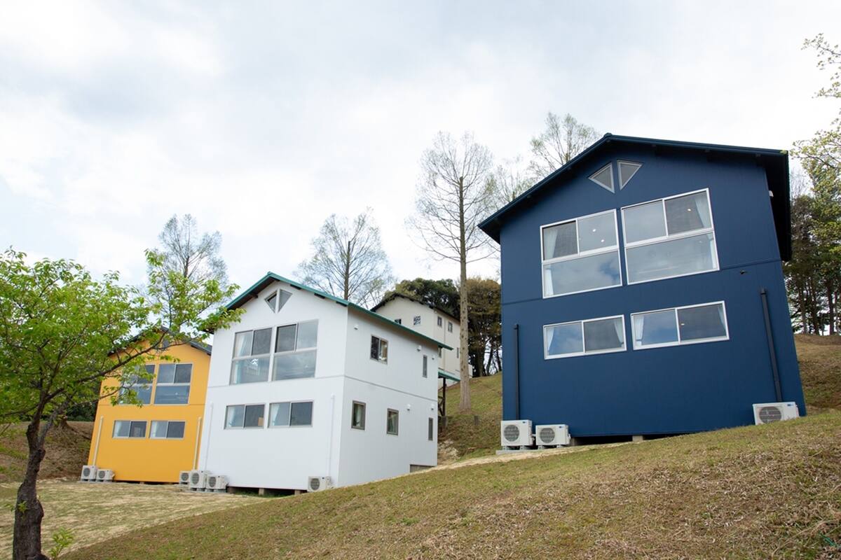 Trois maisons modernes aux couleurs distinctes, situées sur une pente herbeuse.