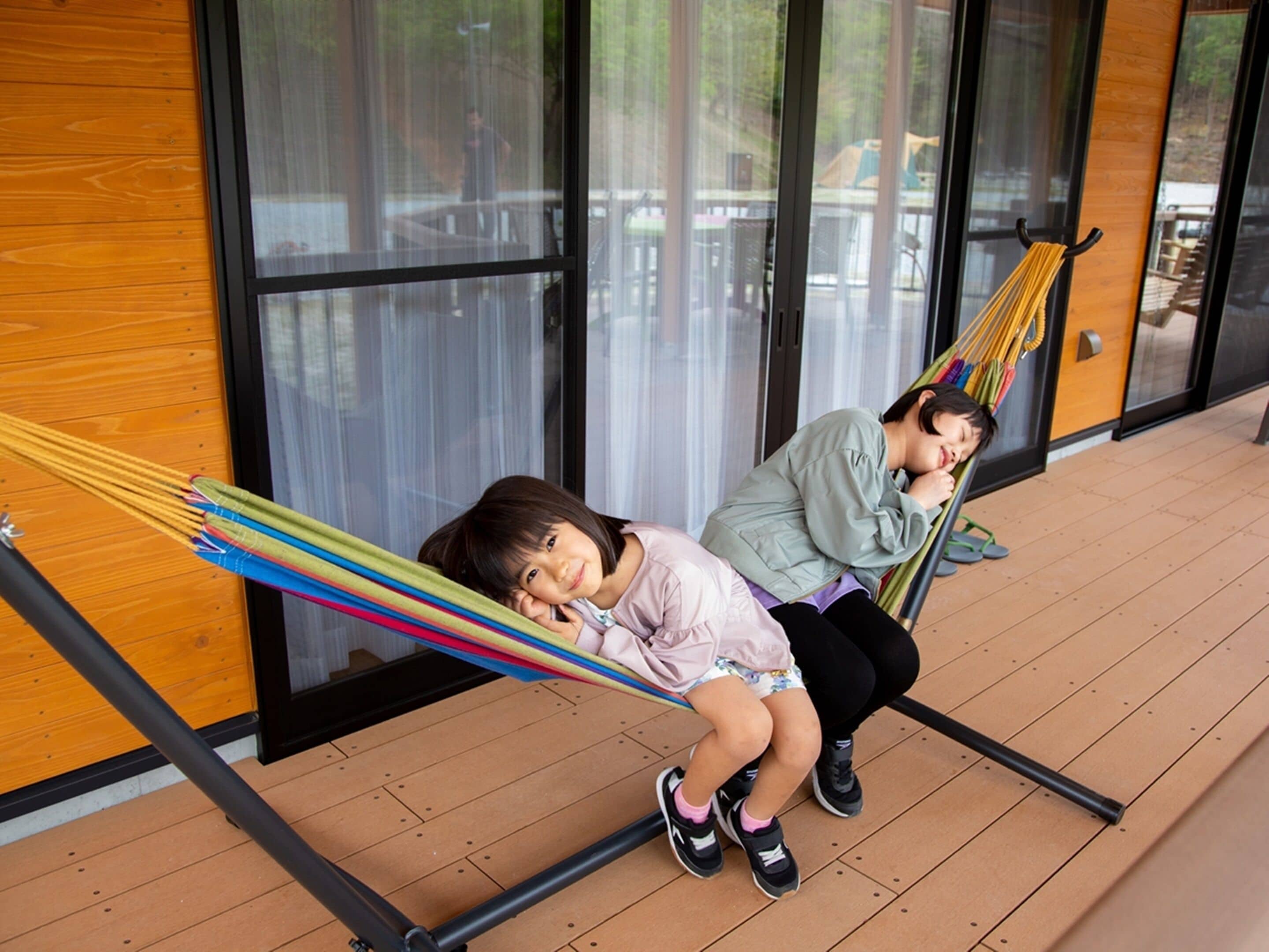 Twee kinderen ontspannen in een hangstoel voor een gebouw met grote ramen en houten vloeren.