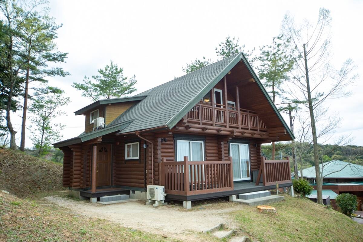 Une cabane en rondins à deux étages, dotée d’un balcon, d’un porche et d’un petit jardin devant.