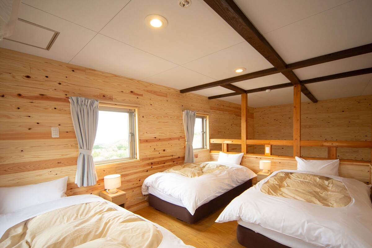 Une chambre dans un chalet en bois, avec trois lits, une fenêtre avec des rideaux et un plafond avec des poutres apparentes.
