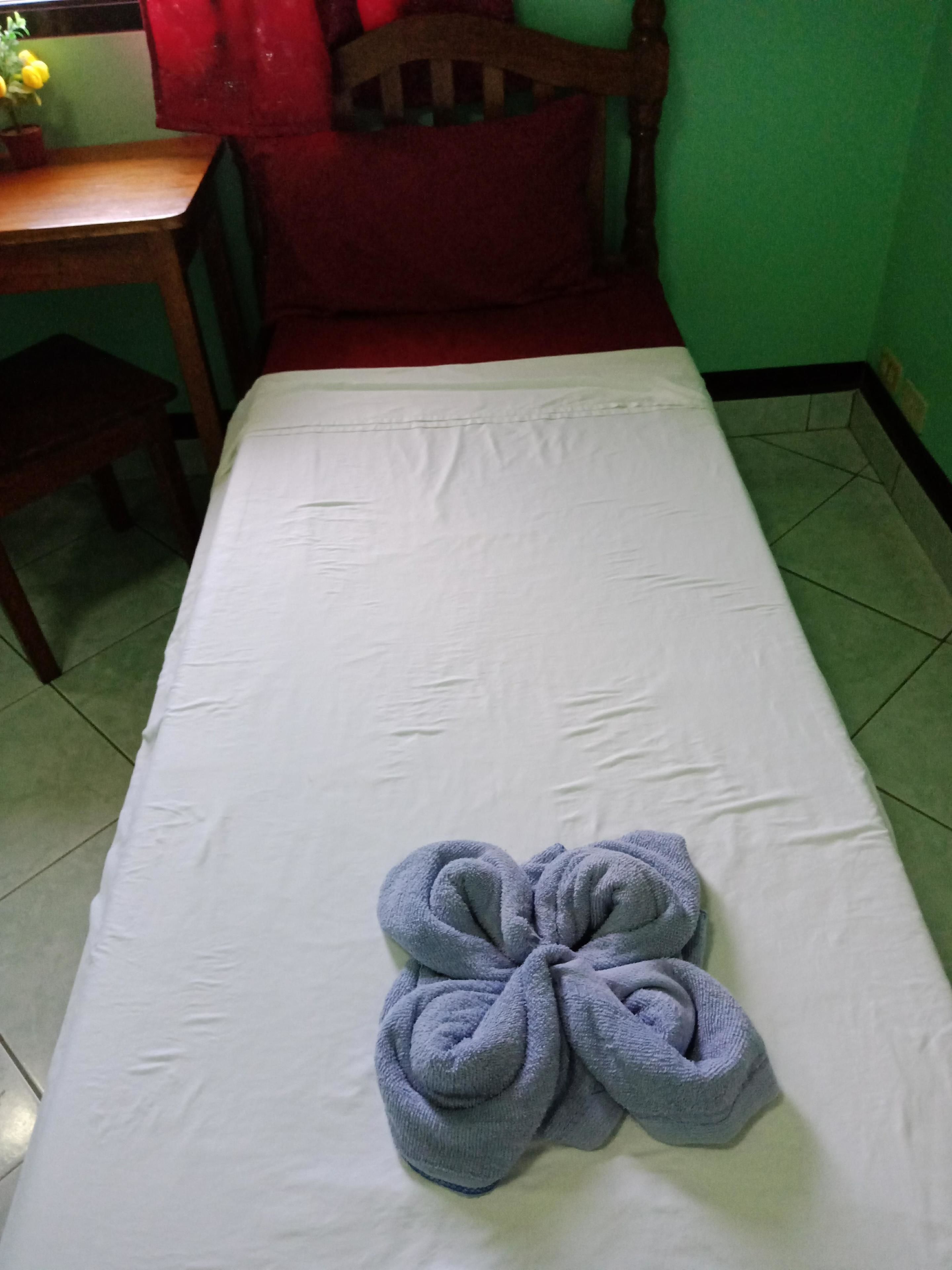 A single bed with a white mattress and a blue towel folded in a flower shape.