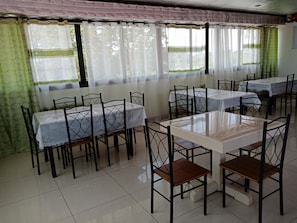 A dining area with tables and chairs, large windows with curtains, and a tiled floor.