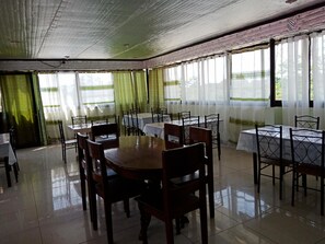 A dining area with tables and chairs, large windows with curtains, and a tiled floor. - Tojo Inn (Puerto Princesa)