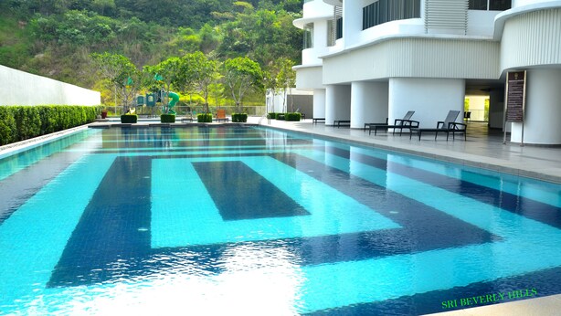 A swimming pool with a clear blue water feature, surrounded by a modern building with a flat roof and a landscaped area with trees and seating. - Sri Beverly Hills Near KLIA & KLIA2 & F1 (Nilai)