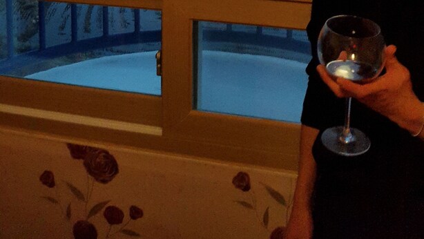 Una persona sosteniendo una copa de vino, mirando un lago con montañas al fondo a través de una ventana con marco decorativo.