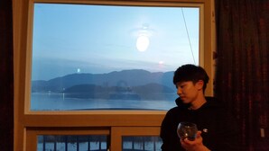 Una persona sosteniendo un vaso junto a una ventana que ofrece vistas a un cuerpo de agua y colinas lejanas.
