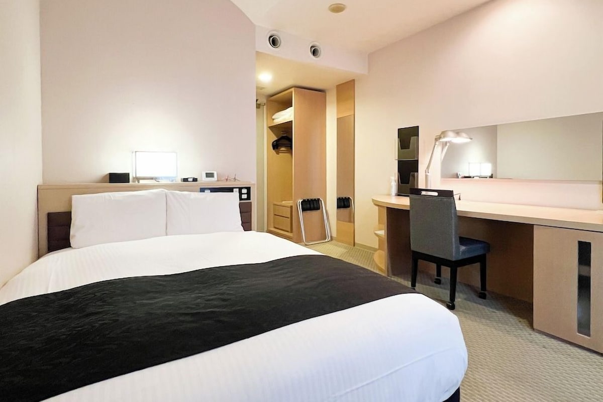 A hotel room with a bed, a desk, a chair, a mirror, and a wardrobe.