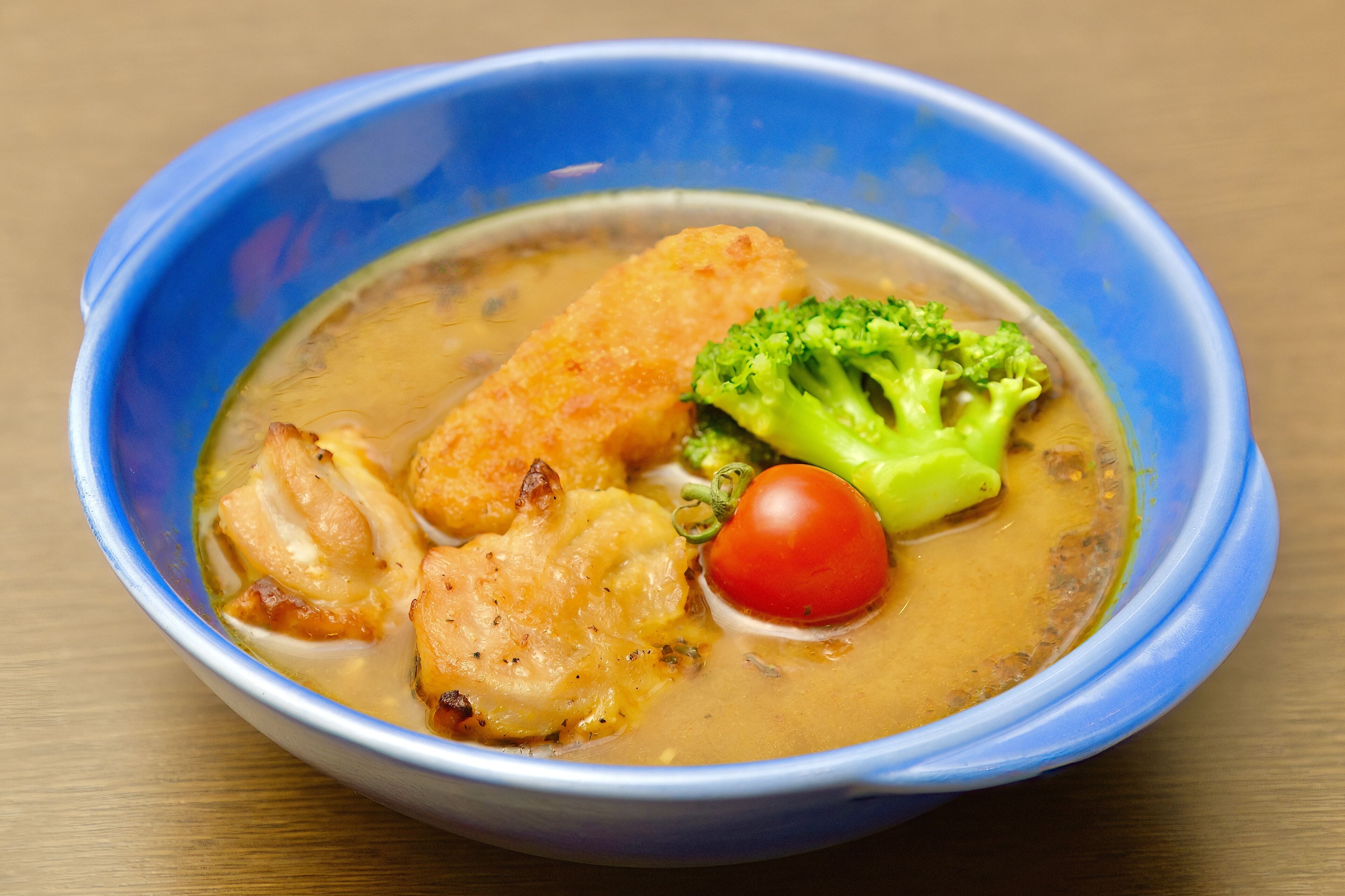 A bowl of soup with chicken, broccoli, and a cherry tomato.