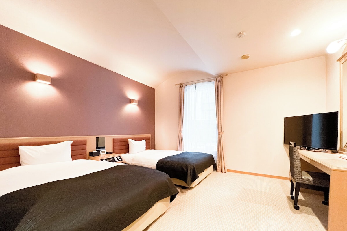Ein Hotelzimmer mit zwei Betten, einem Schreibtisch mit Fernseher und einem Fenster mit Vorhängen.