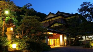 Un edificio japonés tradicional con estructura de madera y un gran acceso, rodeado de árboles e iluminado por farolas.
