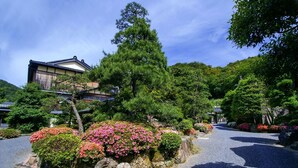 Un edificio japonés tradicional con tejado de tejas, rodeado de abundante follaje y arbustos florecientes.